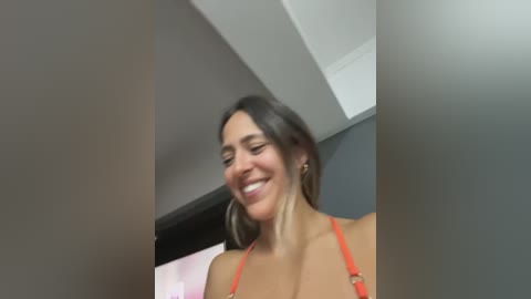 Video of a smiling, tan-skinned woman with shoulder-length dark hair, wearing a red bikini top, standing indoors against a grey wall.