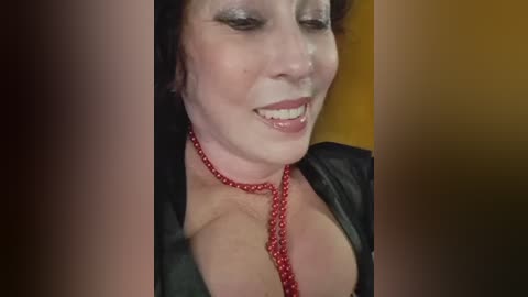 Video of a smiling, fair-skinned woman with short dark hair, wearing a black leather jacket and a red beaded necklace. Background is a blurred yellow and brown gradient.