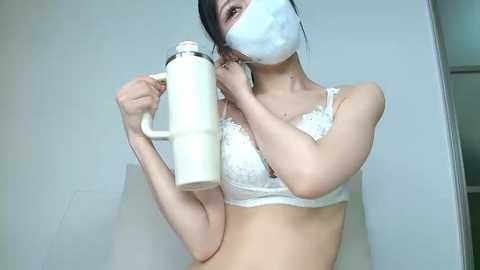 Video of an Asian woman with light skin and black hair, wearing a white lace bra, face mask, and holding a white jug, standing against a plain white wall.