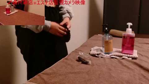 A video of a person wearing a black apron and a grey shirt, standing at a table with a brown tablecloth. On the table are bottles of red and pink liquid, a metal spoon, and a white cloth.