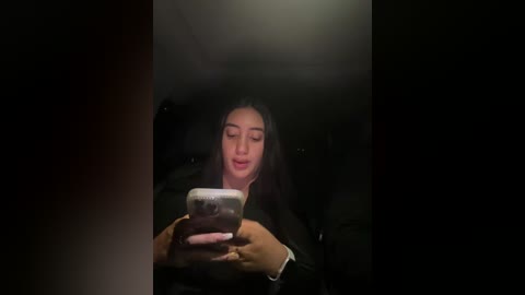 A video shows a woman with long black hair, wearing a dark jacket, smiling while looking at a smartphone in a dimly lit, possibly underground train carriage.