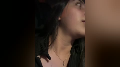 A close-up video of a woman with long black hair, wearing a black dress, with a man's hand on her shoulder, in a dimly lit room.