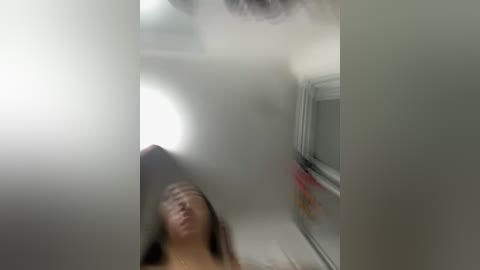 A blurred video of a woman with light skin and long hair, appearing distressed, standing in a small, dimly lit room with white walls and a window on the right.