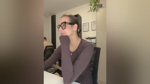 Video of a young woman with glasses, dark hair, wearing a purple top, leaning on her elbow, in a modern office with a green plant, white walls, and framed certificates.