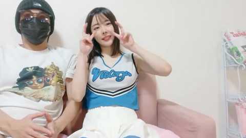 A video of an East Asian woman in a white and blue cheerleader outfit, sitting on a man's lap in a room with a white wall, a magazine rack, and a helmeted man in a white shirt with a cartoon graphic.