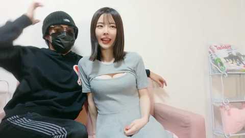 A video of an Asian woman with straight black hair, wearing a gray dress and mask, smiling, standing beside a man in black clothing and mask, both posing cheerfully indoors.