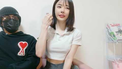 Video of an Asian woman with straight black hair, wearing a white crop top and high-waisted pants, posing in front of a man in a black hoodie with a heart design.