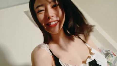 Video of a smiling Asian woman with medium-length dark hair, wearing a black and white lace-trimmed top, standing in a dimly lit room with cream-colored walls.