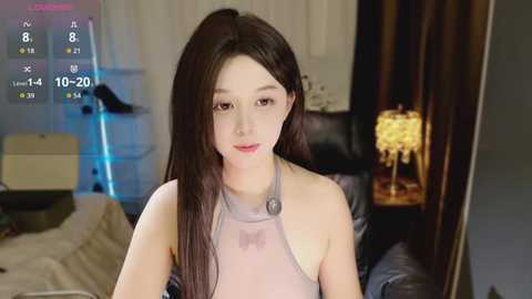 A video of an Asian woman with long black hair in a white halter top, sitting in a dimly lit room with a beige couch and a table lamp.