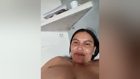 A video of a middle-aged woman with medium skin tone and dark hair, topless, leaning against a white wall with a white shelf holding a white mug and books in the background.
