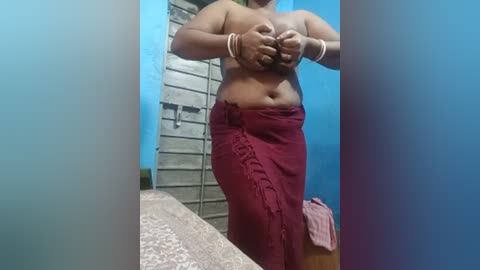 Video of a topless, dark-skinned woman with medium build, wearing a maroon sarong, holding her breasts, standing in a blue-painted room with a tiled shower in the background.