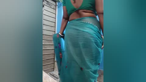 A video shows a woman with medium brown skin, wearing a green, ruffled, see-through sari, partially lifting it to reveal her midriff and a green bra. She stands in a narrow, teal-painted hallway with wooden walls and a metal grate on the floor.