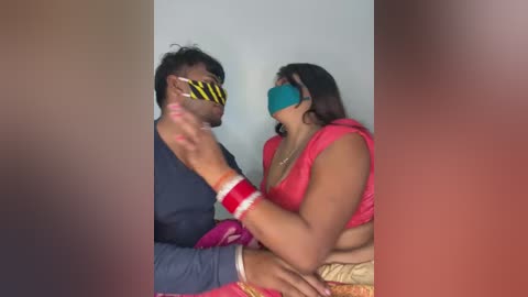 Video of a couple in a bedroom, man in blue shirt, woman in pink sari, both wearing masks, intimate close-up, blurred background.