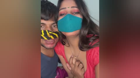 Video of a South Asian couple wearing masks, with the woman's teal mask covering her face and the man's yellow and black mask covering his mouth. They are dressed casually, with the woman in a red top and the man in a blue shirt.