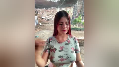 A video of a fair-skinned woman with long, straight red hair, wearing a light green Mickey Mouse t-shirt, standing in an outdoor, rustic setting with a wooden fence and green plants.