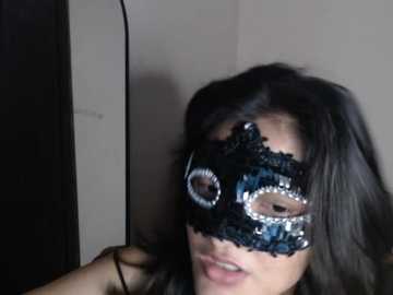Video of a woman with long black hair, wearing a black lace masquerade mask with a silver eye and a black lace headpiece, indoors against a plain beige wall.