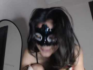 A video of a woman with long black hair wearing a black mask with a lace overlay, grinning widely, standing in a dimly lit room with white walls and a black-framed mirror.