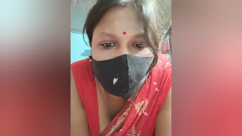 Video of a young woman with a fair complexion, wearing a red saree with intricate gold patterns, black face mask, and red bindi, indoors against a blurred background.