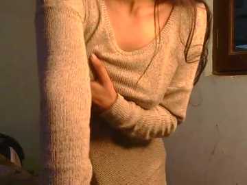 Video of a woman in a beige knit sweater, standing against a beige wall with a window showing a dark room. Her hands are crossed over her chest, and her long brown hair falls over her shoulders.