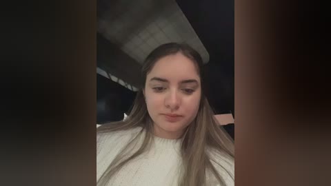 Video of a young woman with light skin and long, straight, brown hair, wearing a white, textured sweater, seated in a dimly lit room with a visible ceiling fan.