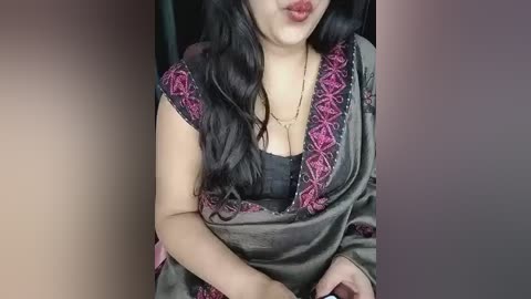 Video of a South Asian woman with long black hair, wearing a black blouse and a green sari with pink floral embroidery. She has a medium complexion and full lips.
