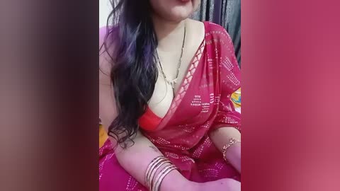 Video of a woman with long black hair, wearing a red sari with gold patterns, exposing a red bra, adorned with gold bangles, sitting on a bed with a pink sheet.