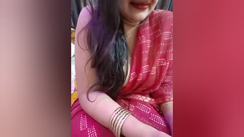 Video of a woman with long black hair in a red, gold-patterned saree, wearing gold bangles. She sits against a red backdrop, smiling slightly.