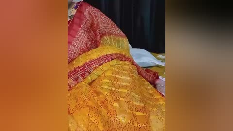 Video of a person lying on a bed, covered in a colorful, patterned blanket with red, yellow, and orange hues. The background shows dark curtains and a white pillow. The scene is warm and cozy.