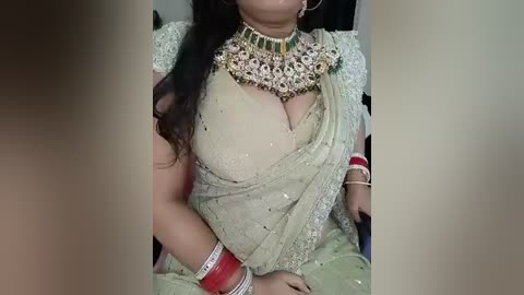 Video of a woman with medium skin tone, wearing a beige saree with intricate gold embroidery, gold jewelry, red bangles, and a large golden necklace. Background is blurred.
