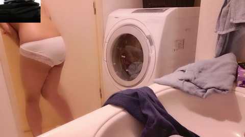 Video of a white woman in a beige bra and panties standing beside a white washing machine in a beige-tiled laundry room, with a blue towel on the washing machine and a black shirt draped over the sink.