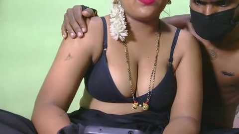 Video of a topless South Asian woman with medium skin tone, wearing a black bra and a floral necklace, partially covered by a man with dark skin, both wearing face masks.