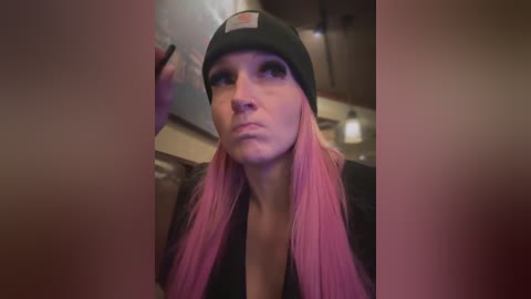 A video of a fair-skinned woman with pink hair, wearing a black beanie and a black top, making a pouty face in a dimly lit indoor setting.