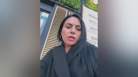 Video of a woman with long black hair, wearing a black coat, standing in front of a building with a wooden shutter and greenery.