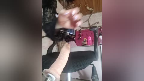 A video shows a person wearing black lace lingerie, with a visible large, erect penis, sitting in a pink chair. The background includes a wooden wall, a pink bag, and a messy desk.
