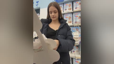 Video of a young girl with straight brown hair, wearing a black puffer jacket, examining a white baby formula box in a brightly lit baby products aisle.