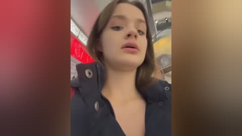 A video of a young woman with fair skin, brown hair, and a dark jacket, standing in a subway car with a red and yellow background.