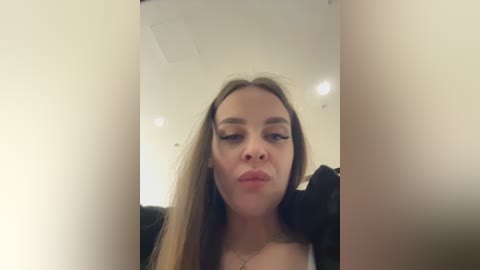 A video of a young woman with long, straight brown hair, light skin, and full lips, captured from a slightly tilted angle. She wears a black top and is indoors with white walls and recessed lighting.