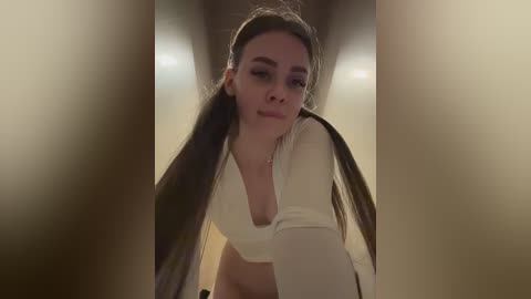 A video of a young woman with long, straight brown hair and fair skin, wearing a white long-sleeve crop top, leaning forward, looking directly at the camera in a dimly lit hallway.