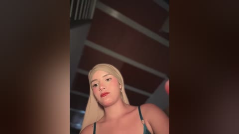 Video of a blonde woman with fair skin, wearing a teal bra, captured from a low angle, showing her cleavage. The background features a dimly lit, modern interior with red and white horizontal beams.