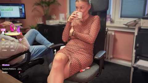 A photo of a blonde woman in a red patterned dress, sitting in an office chair, holding a mug, surrounded by computer monitors, plants, and a desk.