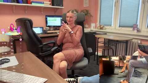 Video of a woman in a patterned dress seated at a desk, using a phone, in a messy office with bookshelves and windows.