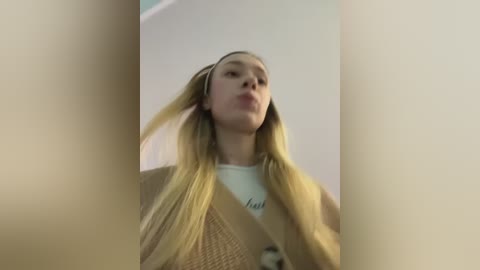A video of a young woman with long blonde hair, wearing a beige jacket and white t-shirt, sticking out her tongue. The background is blurred and light-colored.