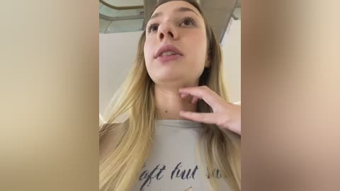 Video of a young woman with fair skin, long blonde hair, and a white T-shirt with \"gift hunt\" text, touching her neck. Background shows a ceiling and beige walls.