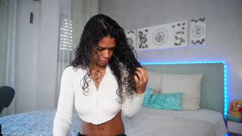 Video of a young woman with long, curly black hair, wearing a white, cropped, long-sleeve top, standing in a modern, minimalistic bedroom with light walls, a bed, and abstract artwork on the wall.