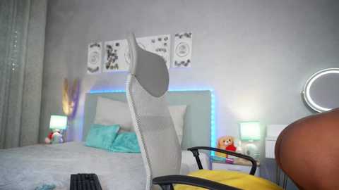 Video of a modern bedroom with a gray upholstered chair, white headboard, turquoise pillows, decorative wall art, and a circular mirror on a white table, lit by soft blue LED lights.