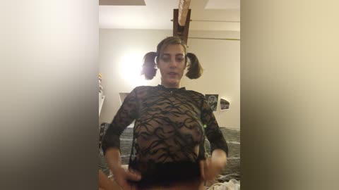 A video of a young woman with light skin, wearing a sheer black top with tiger stripes, and pigtails, standing in a dimly lit bedroom with a bed and minimal decor.