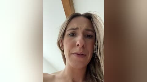 Video of a blonde Caucasian woman with light skin, looking somber, partially obscured by a wooden beam and white curtains in a bright, indoor setting.