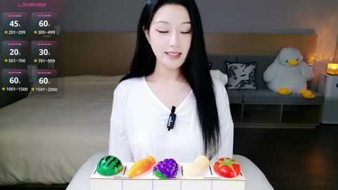 Video of a smiling East Asian woman with long black hair, wearing a white top, holding a colorful fruit tray in a modern bedroom with a gray couch and a plush white duck.