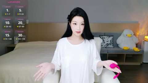 Video of an East Asian woman with long black hair, wearing a white oversized shirt, holding a pink vibrator, in a modern bedroom with a bed, nightstand, and stuffed toys.