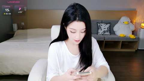 Video of an Asian woman with long black hair, fair skin, and a slender build, wearing a white shirt, sitting on a white sofa, engrossed in a smartphone, in a modern, minimalistic bedroom with a white bed, grey cushions, and a stuffed duck.